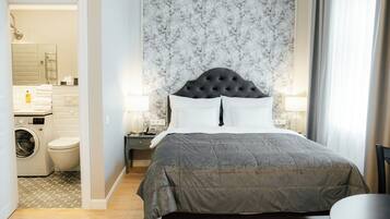 A bedroom with a large bed, a nightstand, a wall-mounted lamp, and a view of a bathroom with a washing machine and toilet.