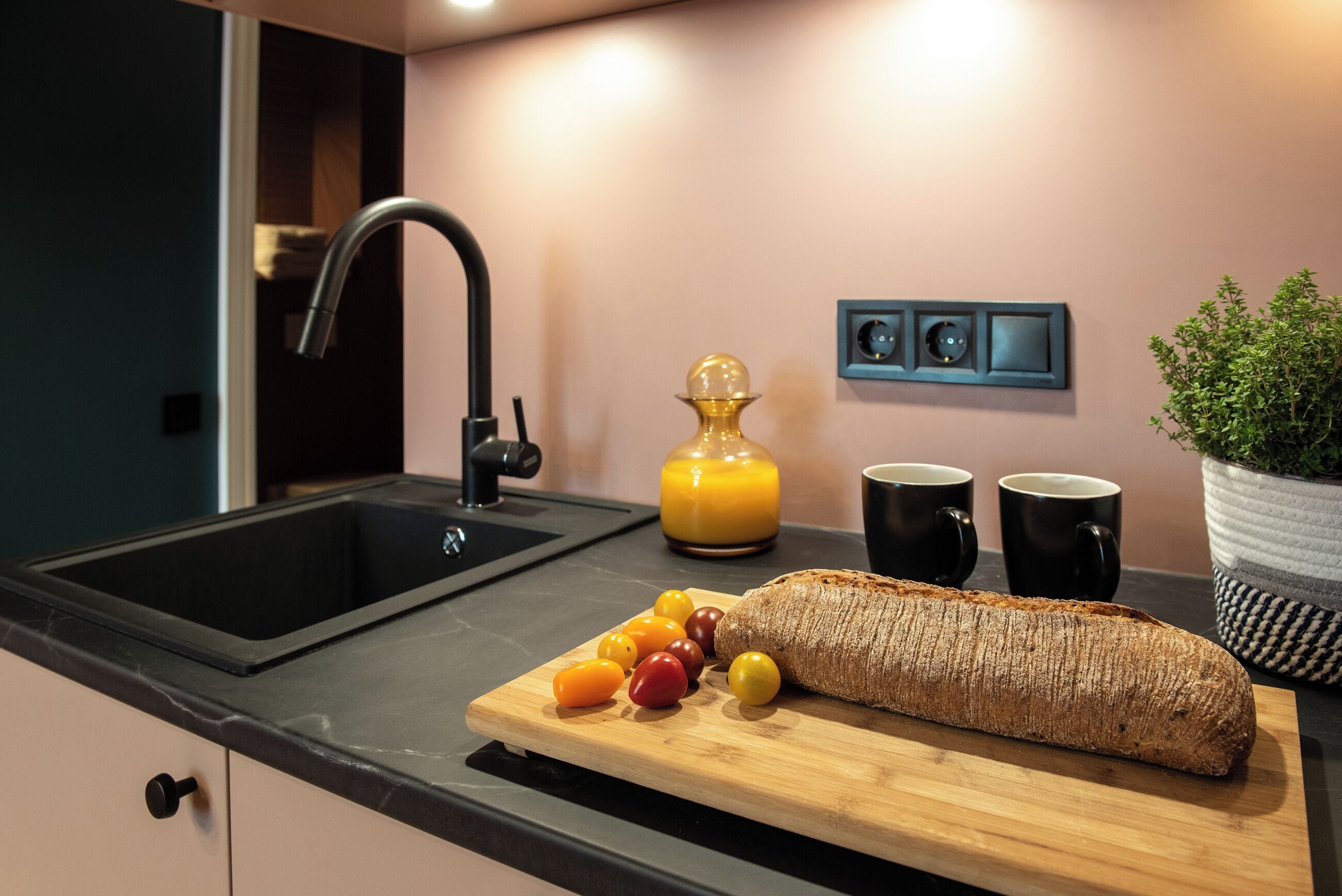 A kitchen with a black sink, a wooden cutting board with tomatoes, a loaf of bread, and two black mugs on the counter.