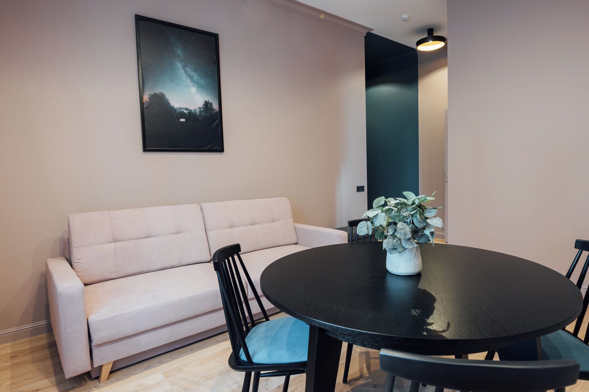 Ein modernes Wohnzimmer mit einem beigen Sofa, einem runden schwarzen Tisch mit Stühlen, einer Zimmerpflanze und einem gerahmten Bild an der Wand.