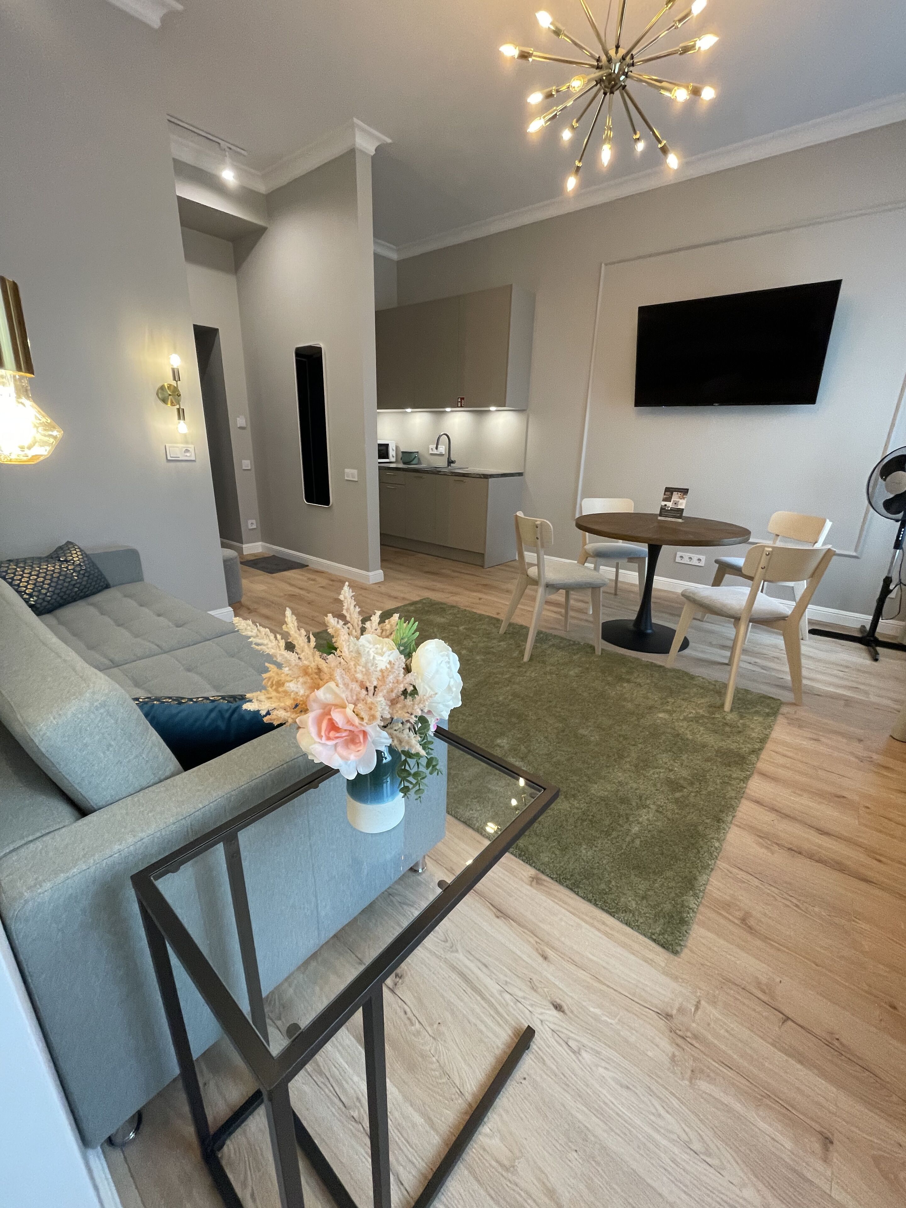 Un salon moderne avec un canapé, une table basse ornée d’un vase de fleurs, un coin repas avec des chaises, une cuisine en arrière-plan et un téléviseur à écran plat fixé au mur.