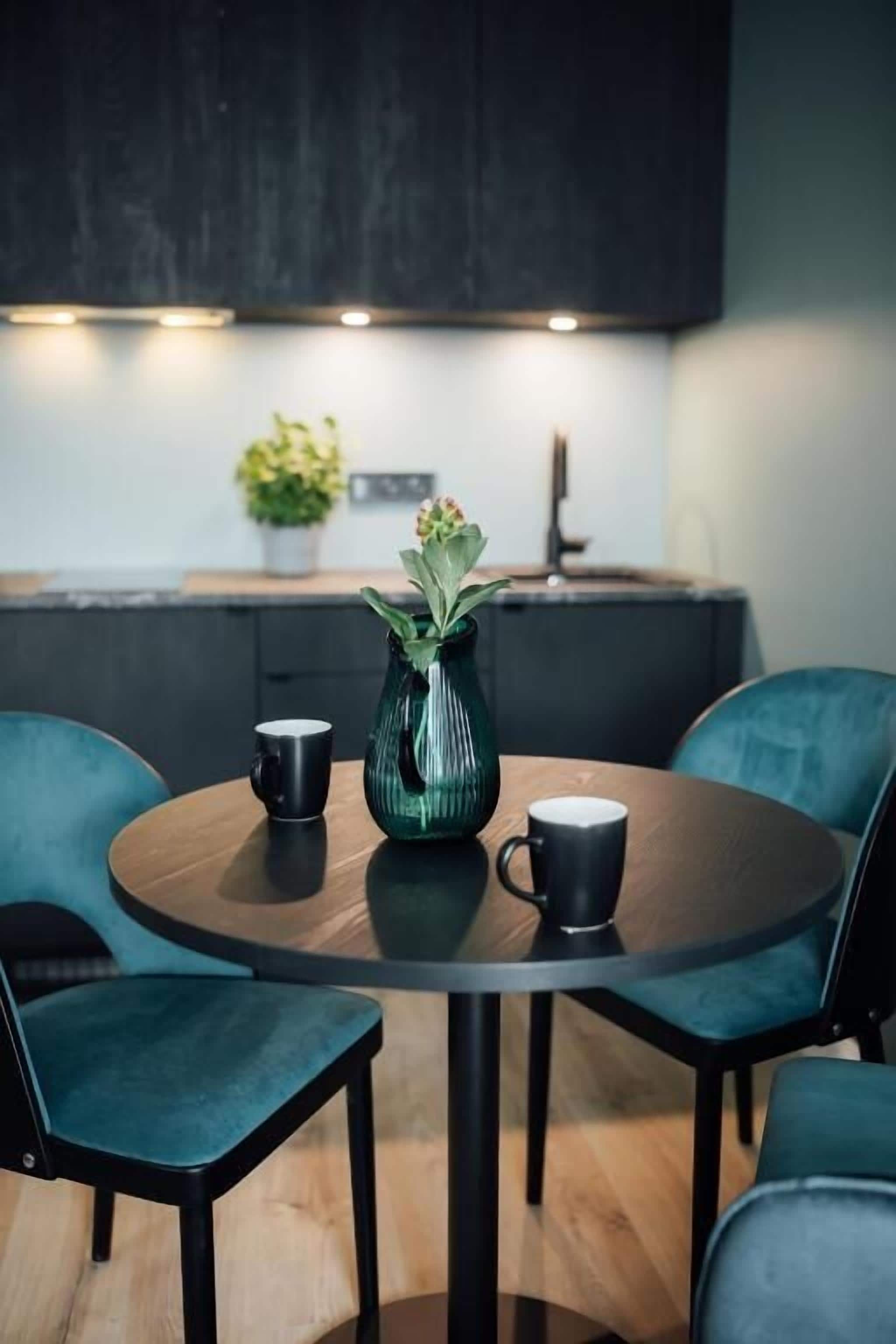 A round table with two black mugs, a green vase with flowers, and two teal chairs in a modern kitchen setting.