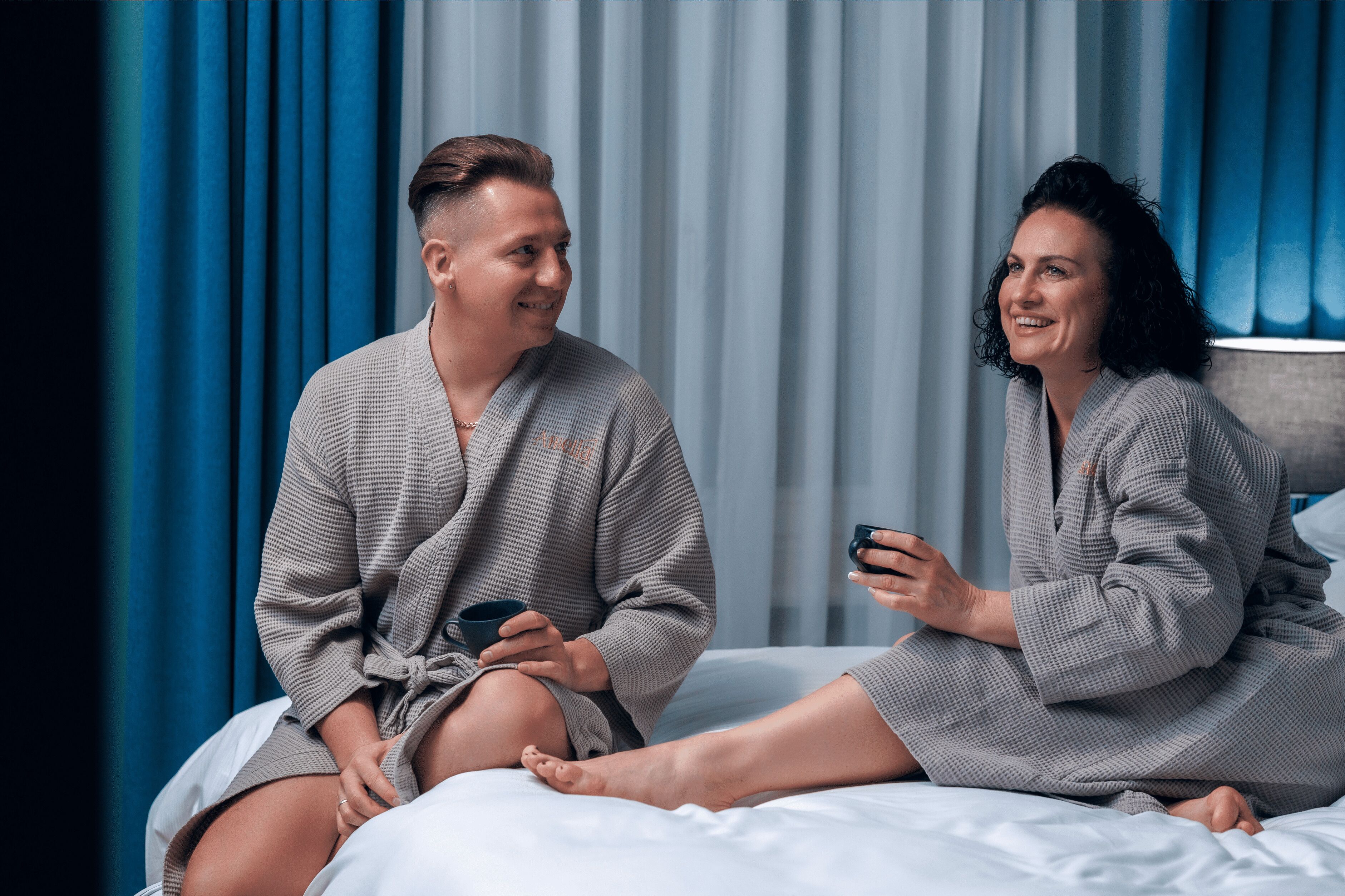 Una pareja con batas a juego sentada en una cama, con vistas a cortinas azules y un cabecero.
