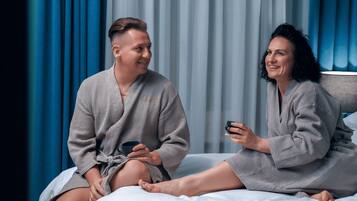 Una pareja con batas a juego sentada en una cama, con vistas a cortinas azules y un cabecero.