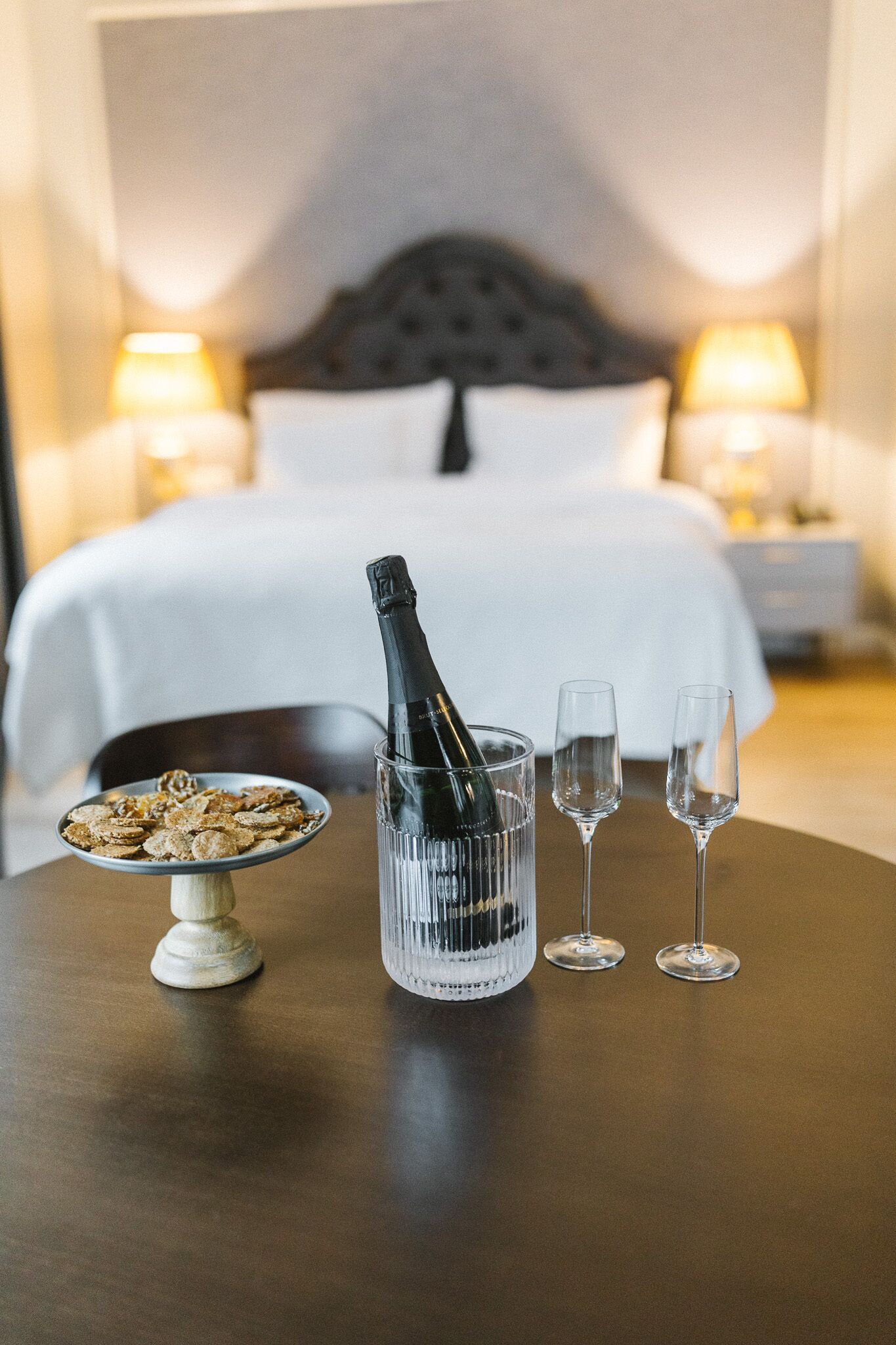 A hotel room with a bed, bedside lamps, a bottle of champagne, two glasses, and a plate of cereal on a table.