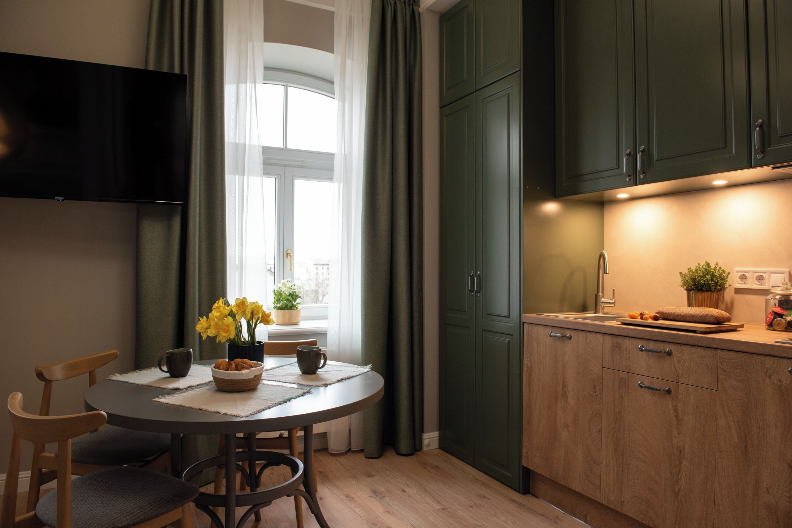 Une cuisine moderne avec des meubles de couleur verte, une table à manger avec des chaises et un téléviseur à écran plat fixé au mur.