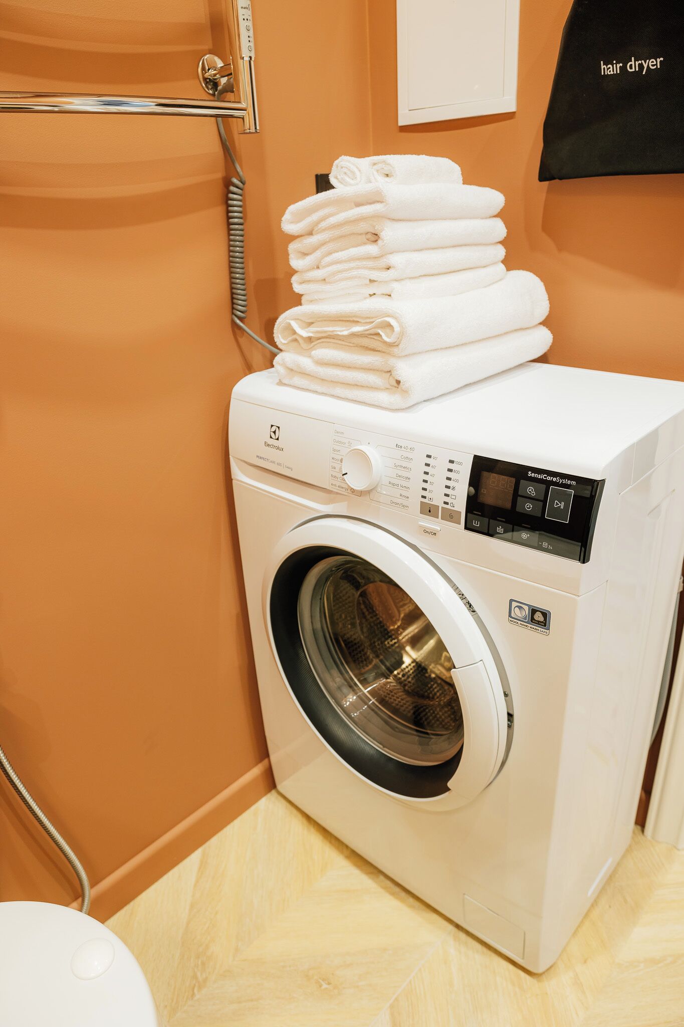A front-loading washing machine with a control panel, stacked towels, and a hair dryer on the wall.