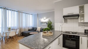 Een moderne keuken met een granieten aanrecht, een ingebouwde oven en witte kastjes. Er is een eethoek met een tafel en stoelen, en een woonkamer met een bank en een kleine salontafel.