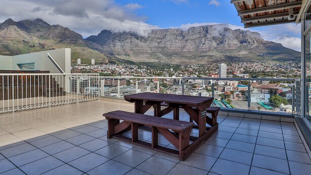 Een balkon met een houten tafel en bankje, met uitzicht op een stadsbeeld en bergen.