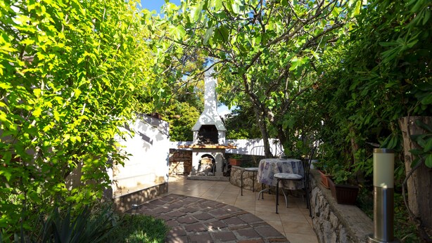 A stone patio with a stone oven, surrounded by greenery and a white wall.