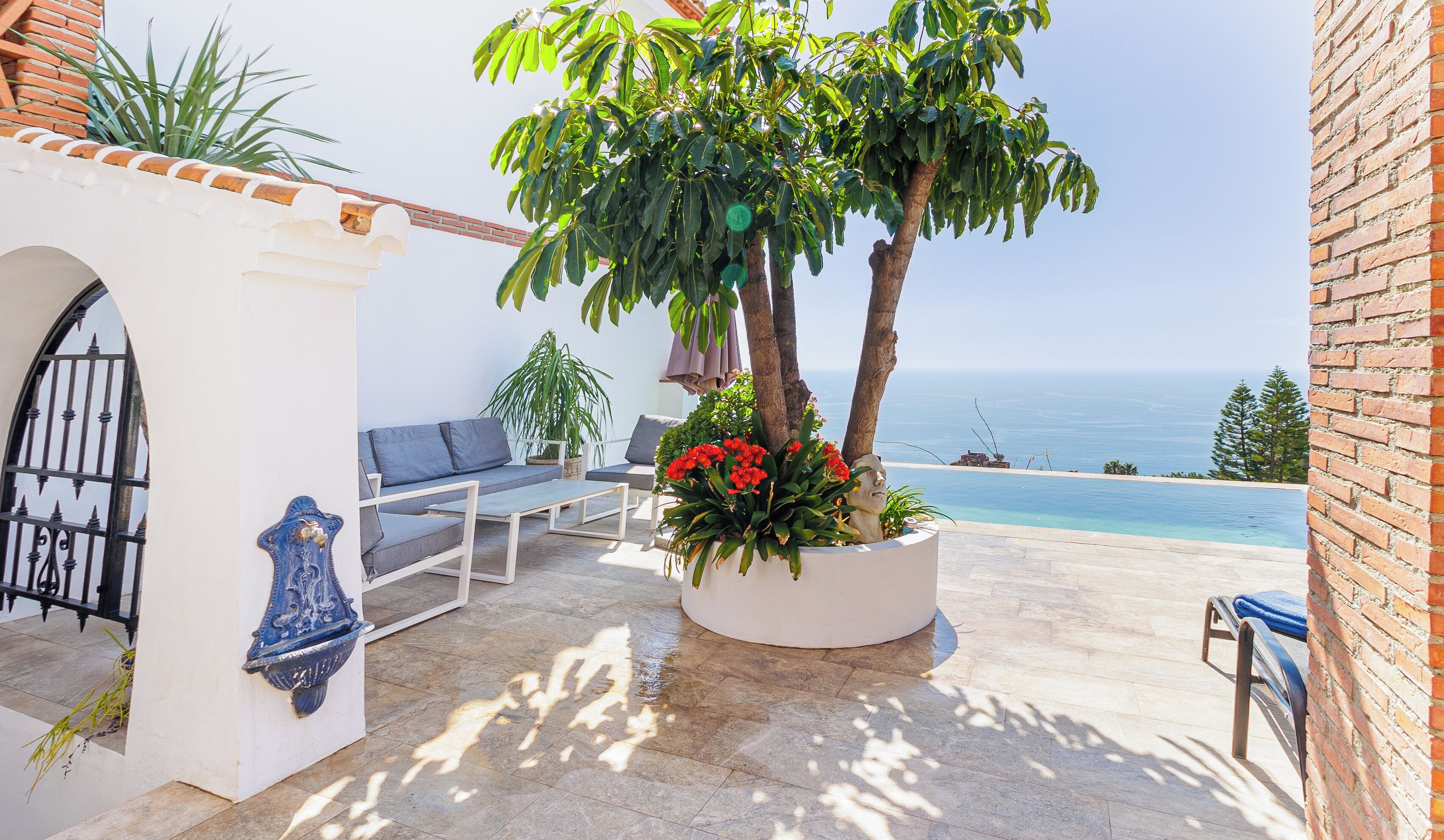 Um pátio com piscina, uma parede branca com uma bacia azul decorativa e uma área de estar com vista para o mar.