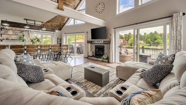 A spacious living room with a high ceiling, a fireplace, and a large window overlooking a balcony. - Great Lake Escape by PMR, 8BR Lakefront, Private Dock & Sun Deck, Sleeps 24 (Lake Harmony)
