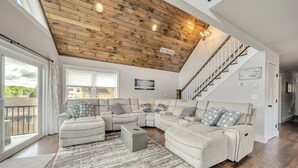 A spacious living room with a wooden ceiling, a large sectional sofa, a grey ottoman, and a view of the outdoors through large windows. - Great Lake Escape by PMR, 8BR Lakefront, Private Dock & Sun Deck, Sleeps 24 (Lake Harmony)