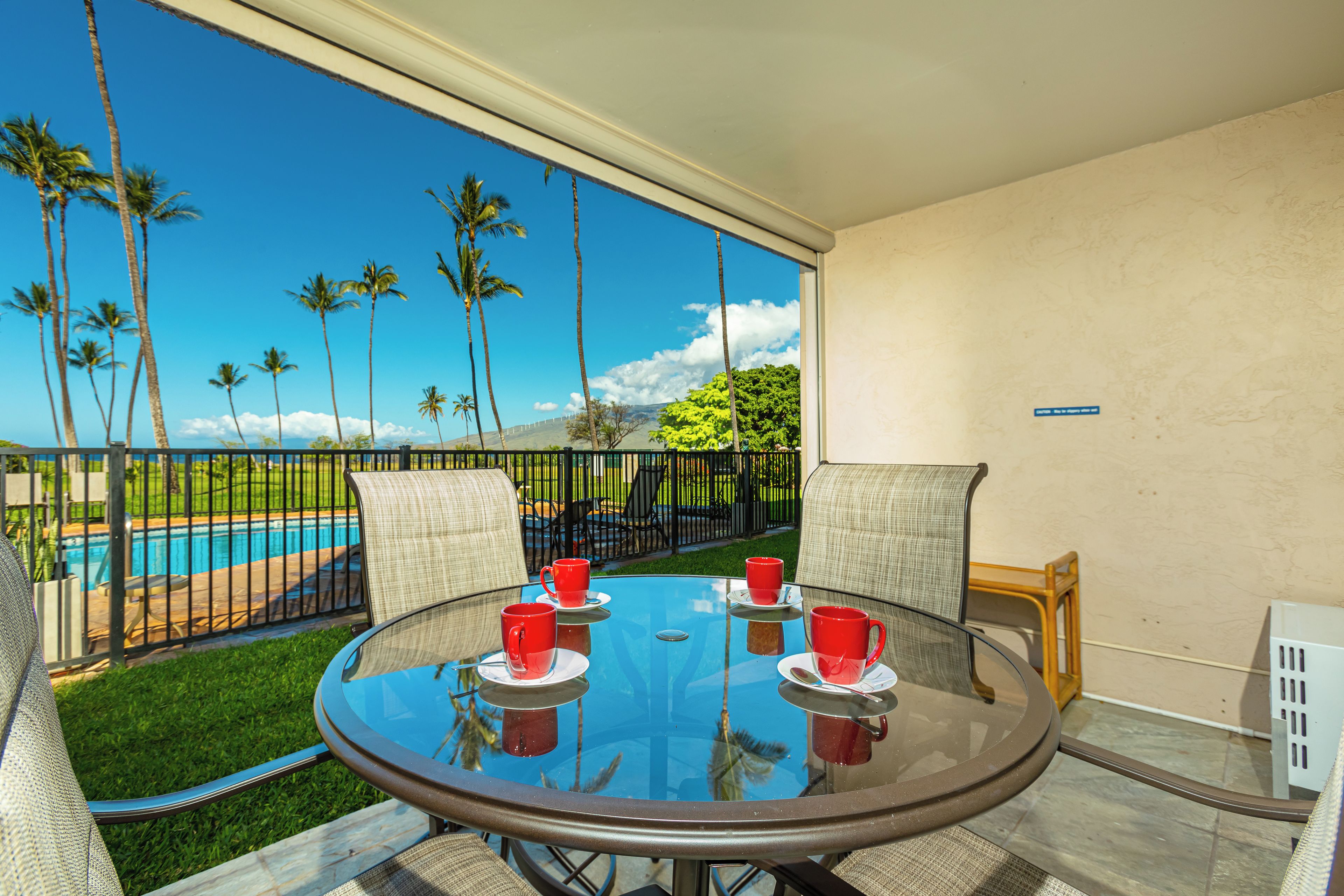 A round glass table with four red cups and saucers, surrounded by wicker chairs, overlooking a swimming pool and palm trees.