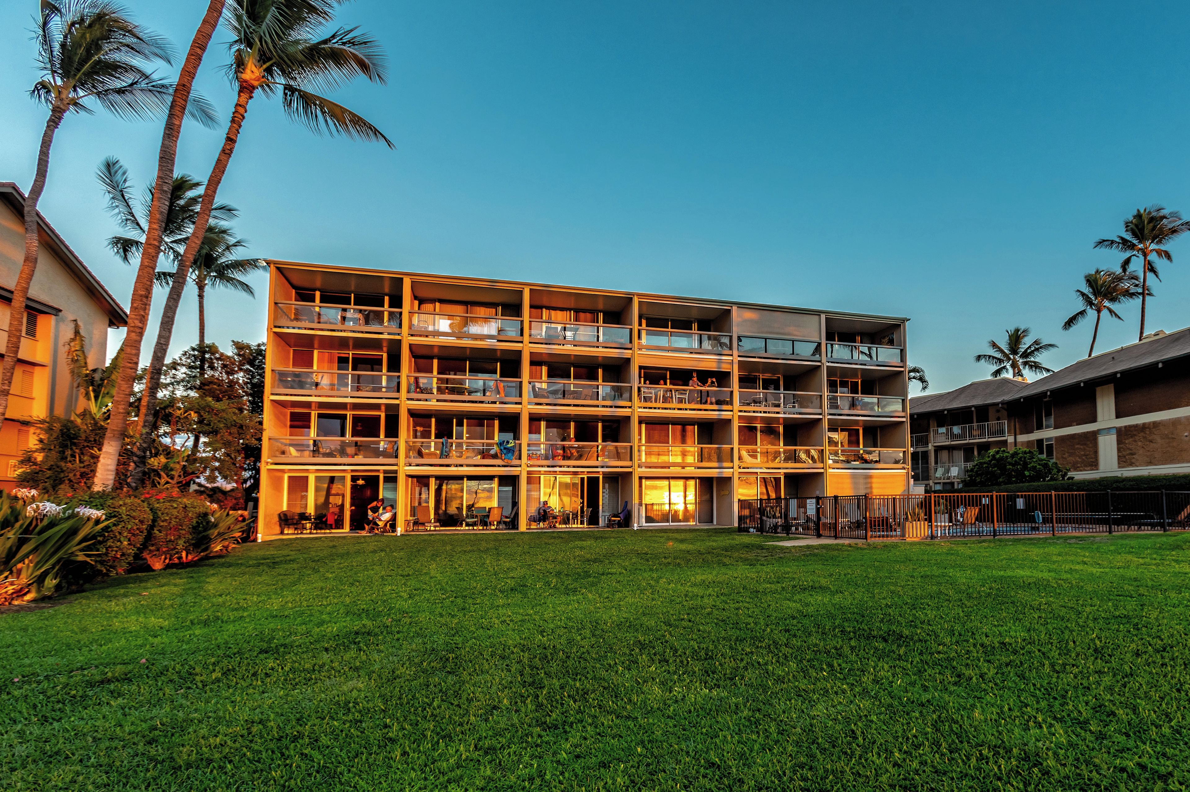 A multi-story hotel with balconies, a grassy lawn, and palm trees.