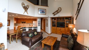 A spacious living area with a brown leather sofa, a wooden coffee table, and a dining table with chairs. There is a kitchen in the background with wooden cabinets and a built-in oven.
