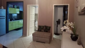 Een woonkamer met een beige bank, een bijzettafeltje met witte bloemen en een keuken die door een open deur zichtbaar is.