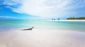 A beach with clear turquoise water, a driftwood piece on the sand, and a few people in the distance.