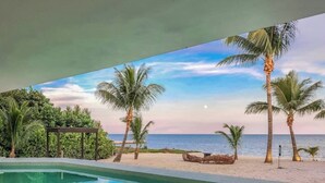 A swimming pool with a view of the beach and ocean, surrounded by palm trees and a gazebo.
