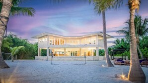 A two-story beachfront property with a large balcony, palm trees, and a sandy beach.