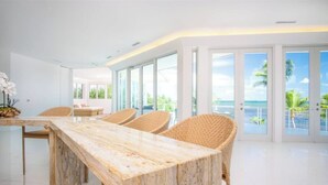 A modern dining area with a marble table and wicker chairs, large windows, and a view of the ocean.