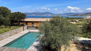 A swimming pool with a wooden deck, surrounded by olive trees and a view of the sea and distant mountains.