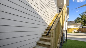 Un escalier en bois, doté d’une rampe métallique, mène à un bâtiment dont le revêtement extérieur est blanc.