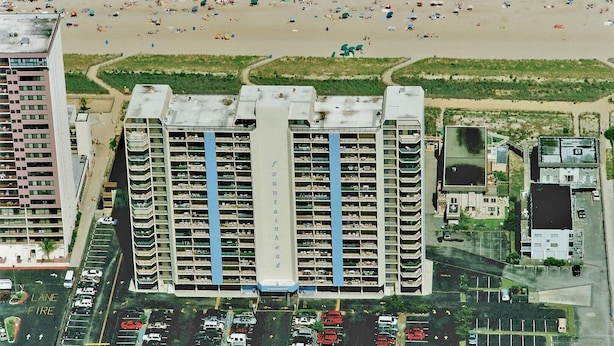 Um hotel moderno alto com vários andares, uma faixa central azul e um estacionamento com diversos carros.