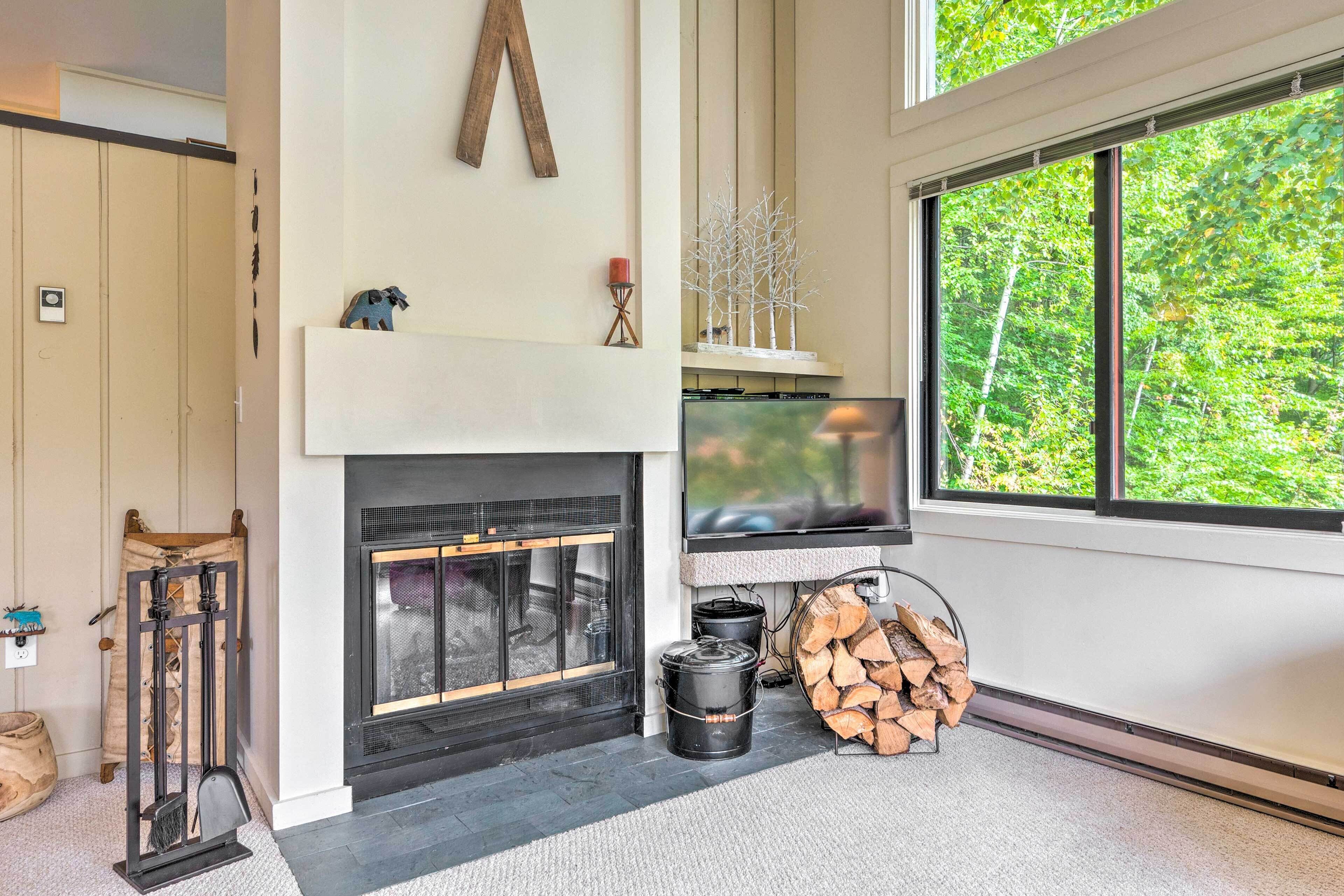 A modern living room with a fireplace, a large window with a view of trees, and a mounted television.
