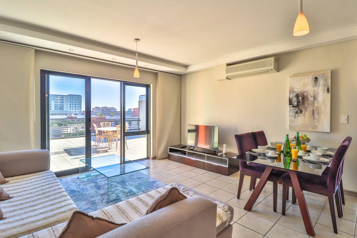 A modern living room with a sofa, a dining table set for two, and a large window offering a view of the city.