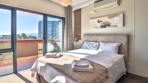 Une chambre avec un grand lit, un balcon donnant sur des immeubles et un climatiseur fixé au mur.