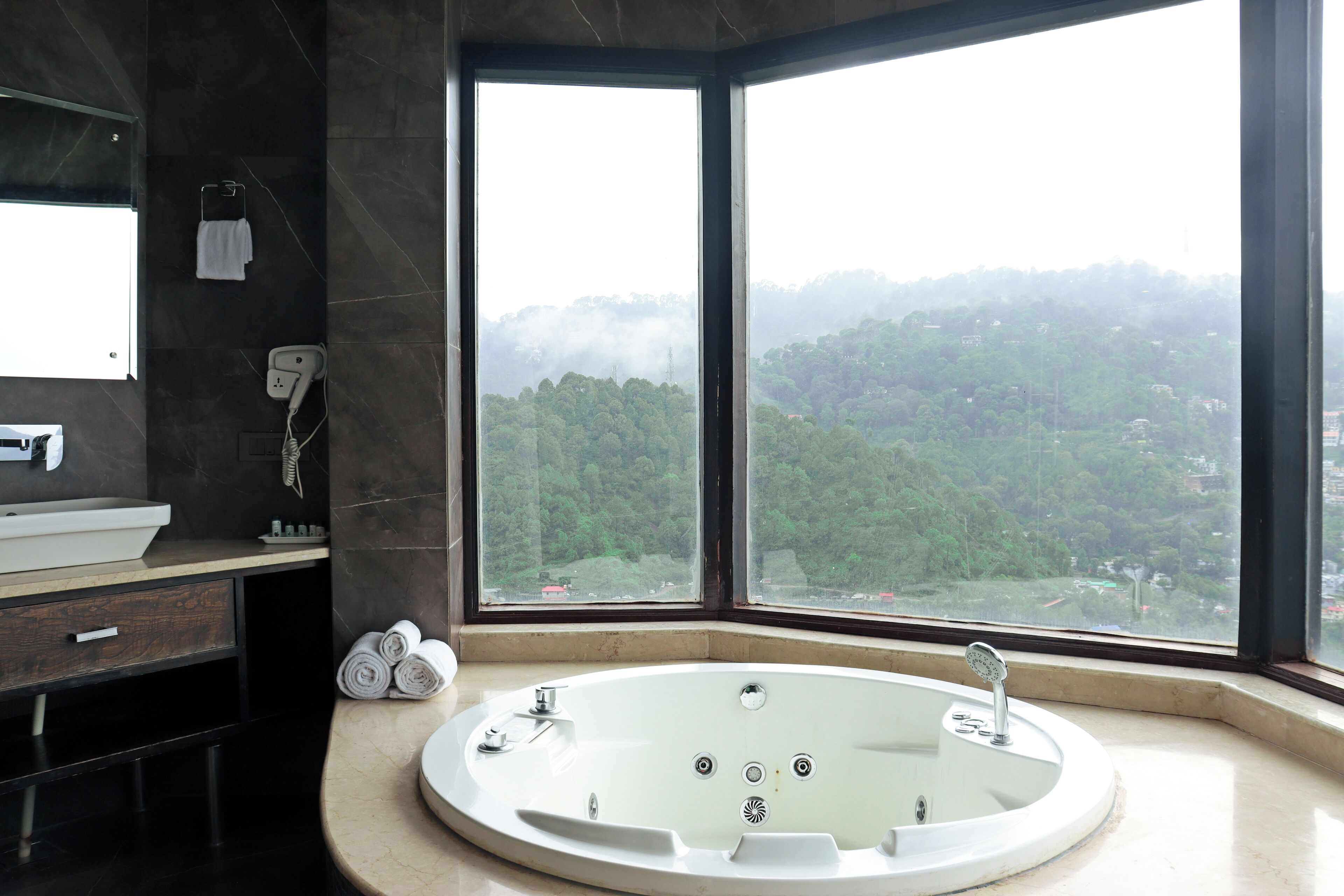 Une salle de bain moderne dotée d’une grande baignoire, d’un meuble-lavabo avec miroir et offrant une vue sur une zone boisée grâce à de grandes fenêtres.