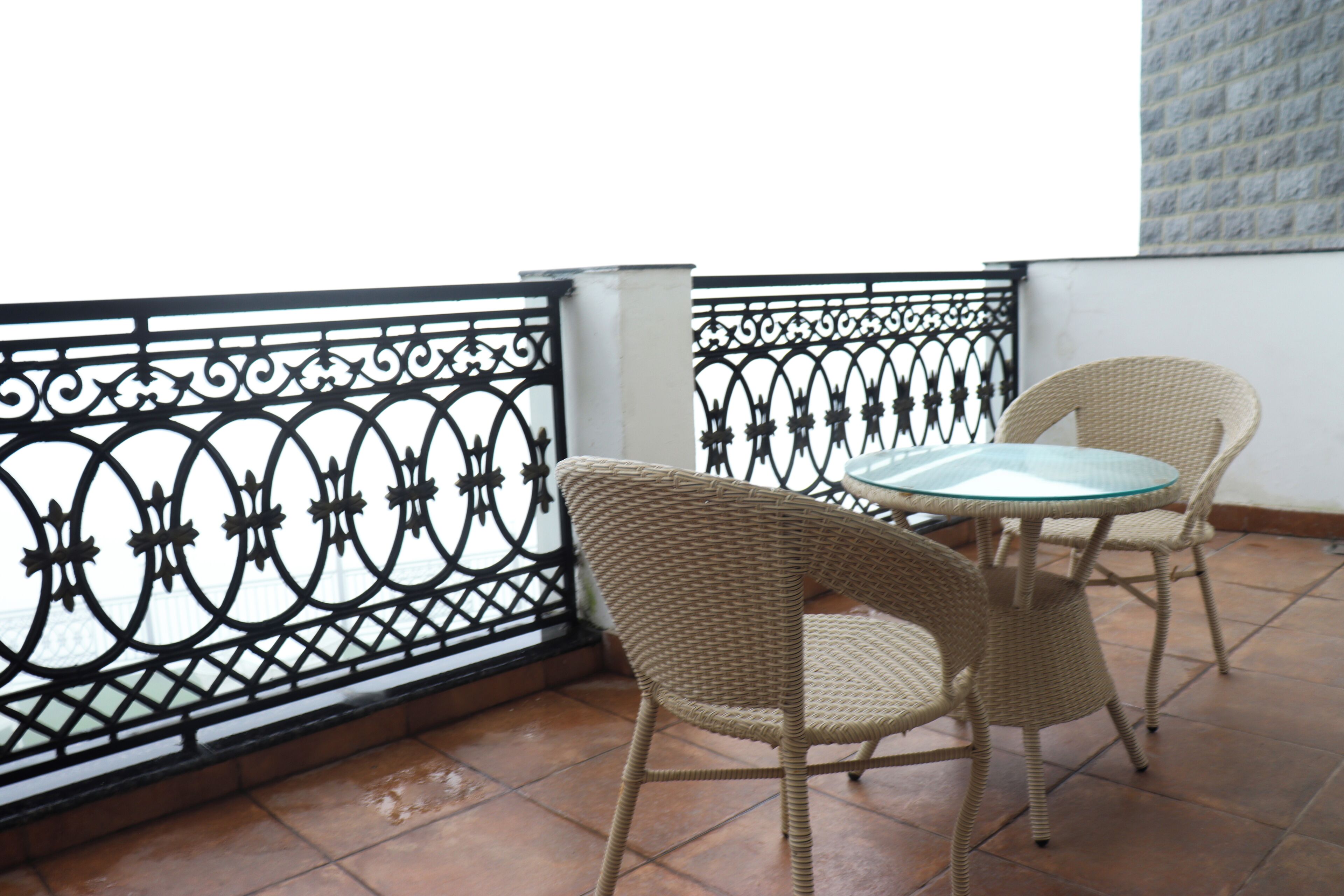 A balcony with a wrought iron railing, two wicker chairs, and a glass table.