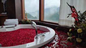 A bathroom with a bathtub filled with rose petals, a window with a view of mountains, and a flower arrangement on the windowsill. - Rosetum Kasauli (Kasauli)