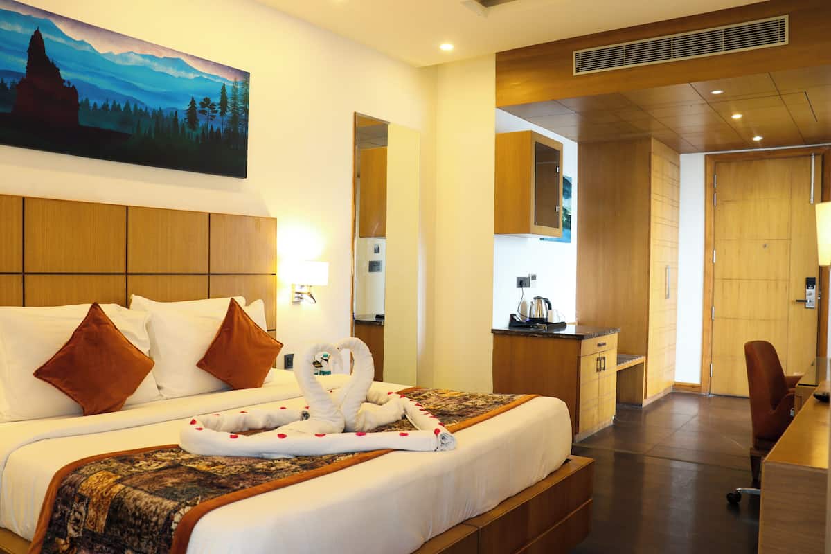 A hotel room with a large bed, a painting of a mountain landscape, a desk with a kettle, and a wooden cabinet.