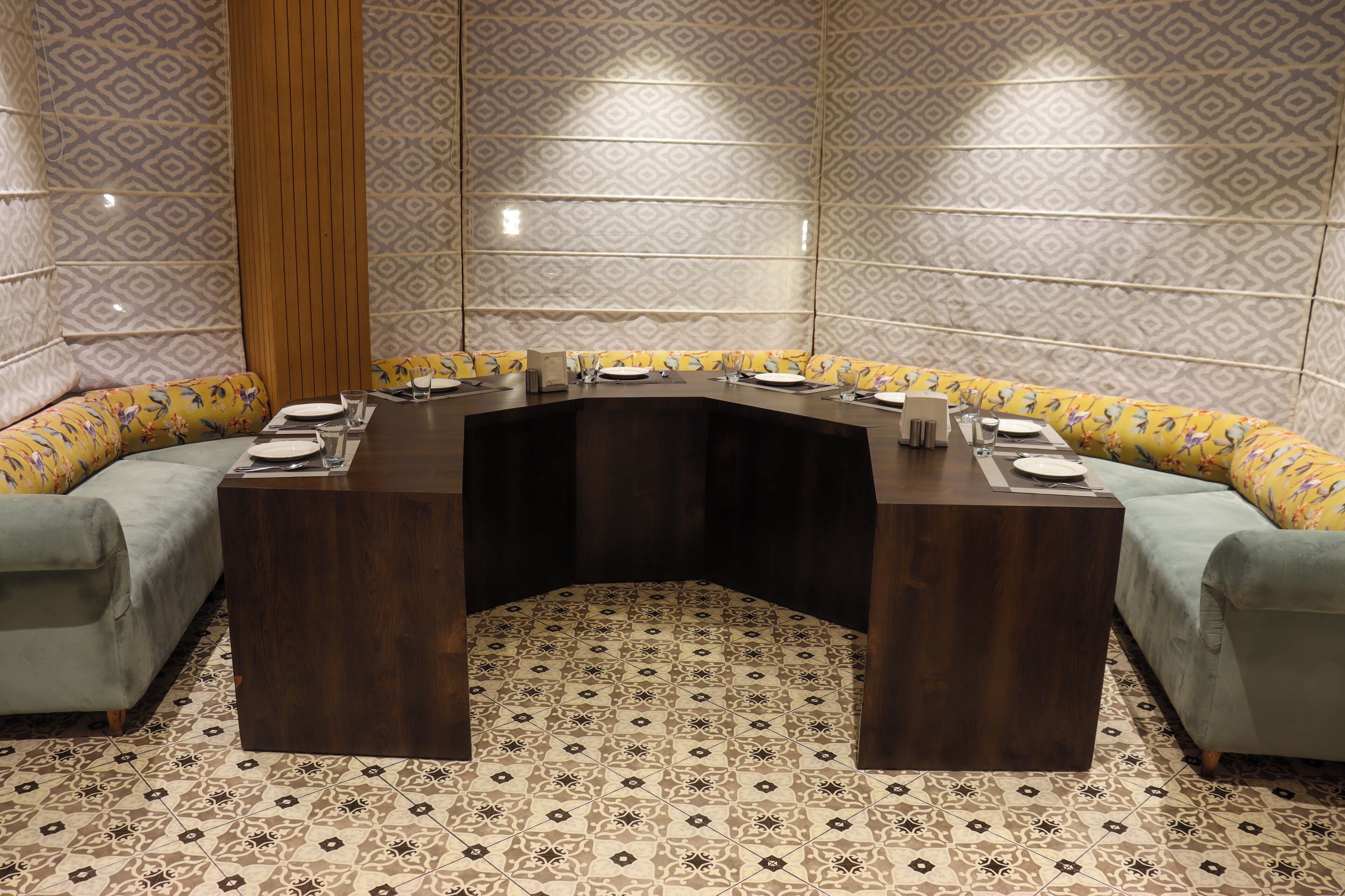 A conference room with a curved dark wood table, light grey sofas with floral upholstery, and patterned floor tiles.