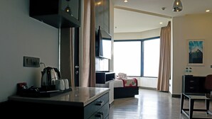 Une chambre d’hôtel moderne équipée d’une kitchenette, d’une grande fenêtre avec des rideaux, d’un lit et d’un tableau au mur.