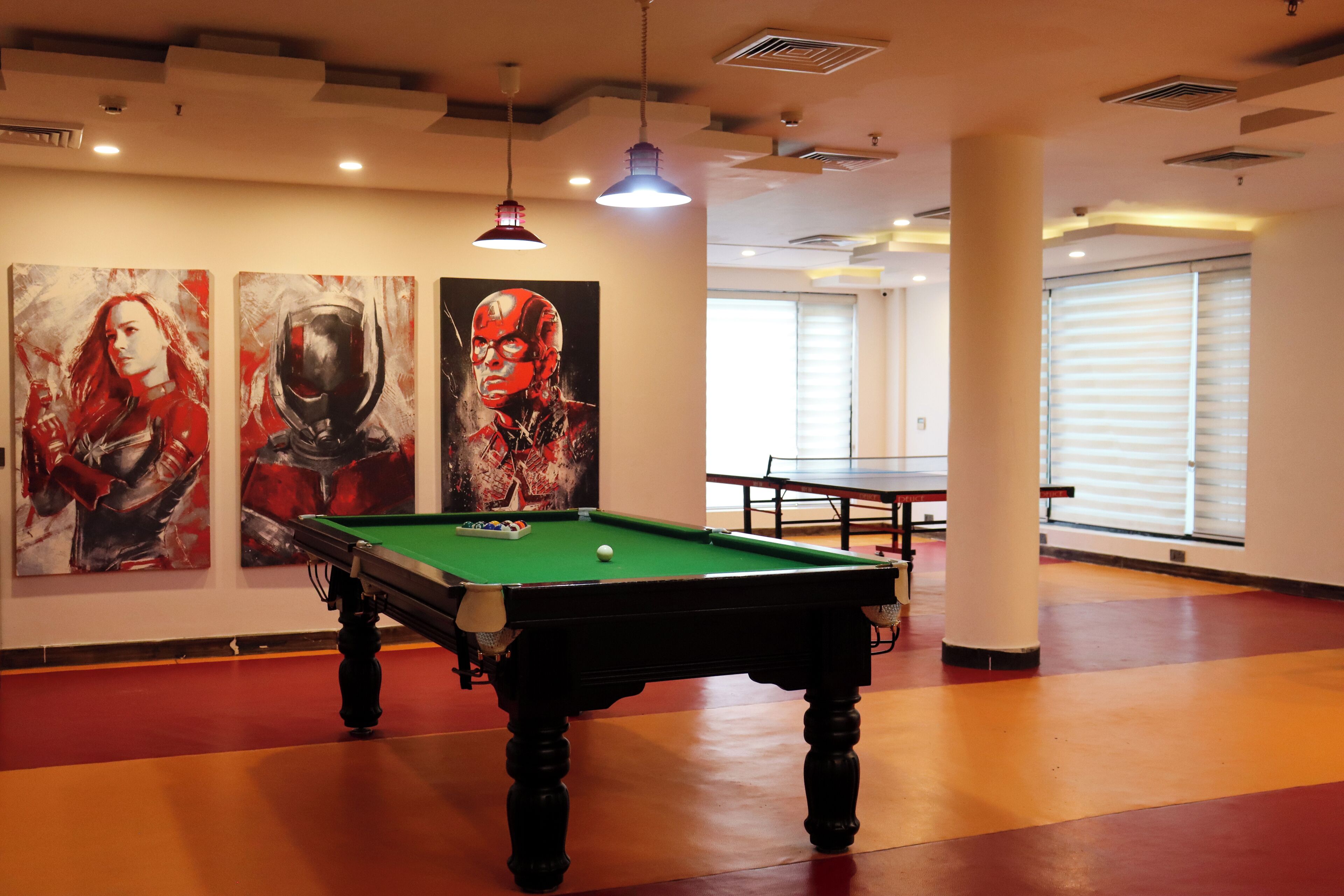 Una mesa de billar en una habitación con tres grandes cuadros en la pared, una mesa de ping pong y un ventanal con persianas.