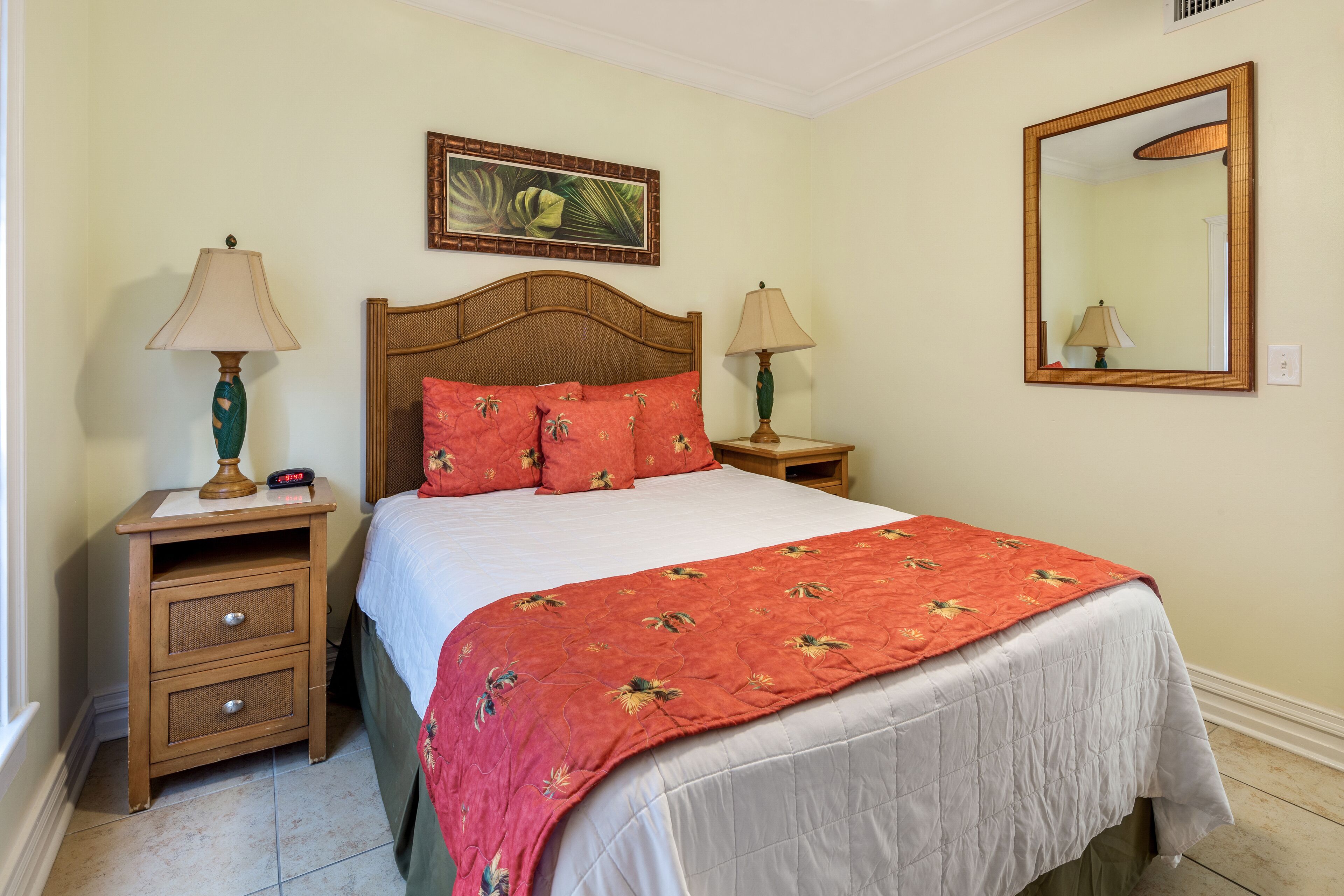 A neatly made bed with a floral bedspread, two bedside tables with lamps, a wall-mounted mirror, and a tropical-themed painting above the bed.