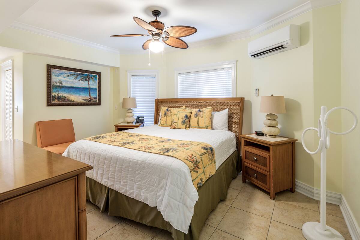 Un dormitorio con cama, escritorio, silla, ventilador de techo, mesita de noche, lámpara y aire acondicionado instalado en la pared.
