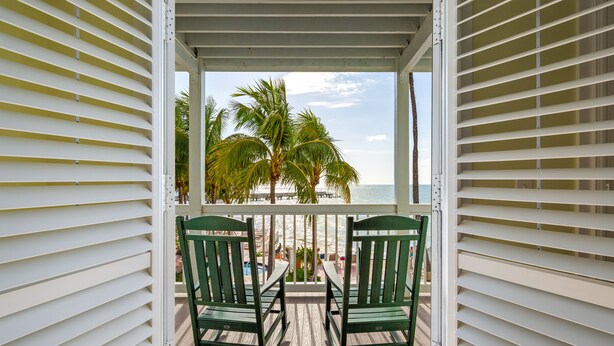 A balcony with two rocking chairs facing a beach with palm trees and a pier.