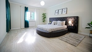 A bedroom with a curved bed, a bedside table with a lamp, a potted plant, and a view of the outside through a window. - Holiday Home Villa As Cumadres with Mountain View, Pool, Garden & Wi-Fi (Azeitão)