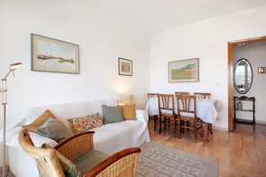 A living room with a white sofa, wicker chairs, a dining table with chairs, and framed artwork on the walls.