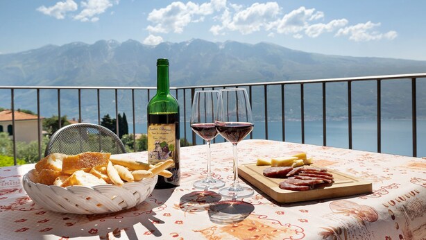 A table with a bottle of wine, glasses, bread, and a board with meats, set against a scenic mountain backdrop. - Holiday Apartment 'Rustico Spagnol 4' with Lake View, Pool & Wi-Fi (Tignale)