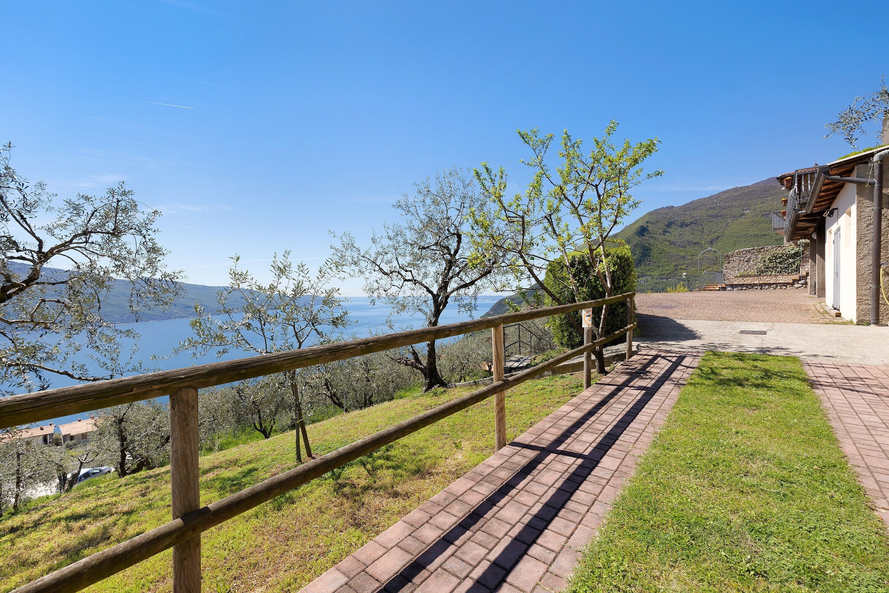 Un sentier pavé bordé d’une rambarde en bois, un bâtiment aux murs de pierre et une vue sur un lac et des montagnes.
