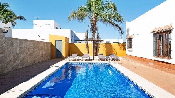 A swimming pool with lounge chairs and a palm tree, surrounded by white and yellow buildings. - Vacation Home "Casa La Vigía" with Private Pool, Private Terraces & Wi-Fi (Conil de la Frontera)
