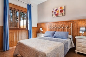 Une chambre à coucher avec une tête de lit en bois à panneaux, un lit avec une literie à motifs, une table de chevet avec une lampe et une fenêtre avec des rideaux bleus.