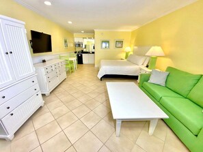 A hotel room with a bed, a green sofa, a white dresser, a white table, and a kitchenette in the background.