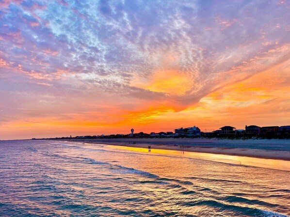A beach with a sunset, calm sea, and buildings in the background.