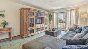 A living room with a sofa, a wooden cabinet with books, a television, and a balcony with curtains. - One Bedroom - Bluewater Villas 109A (Myrtle Beach)