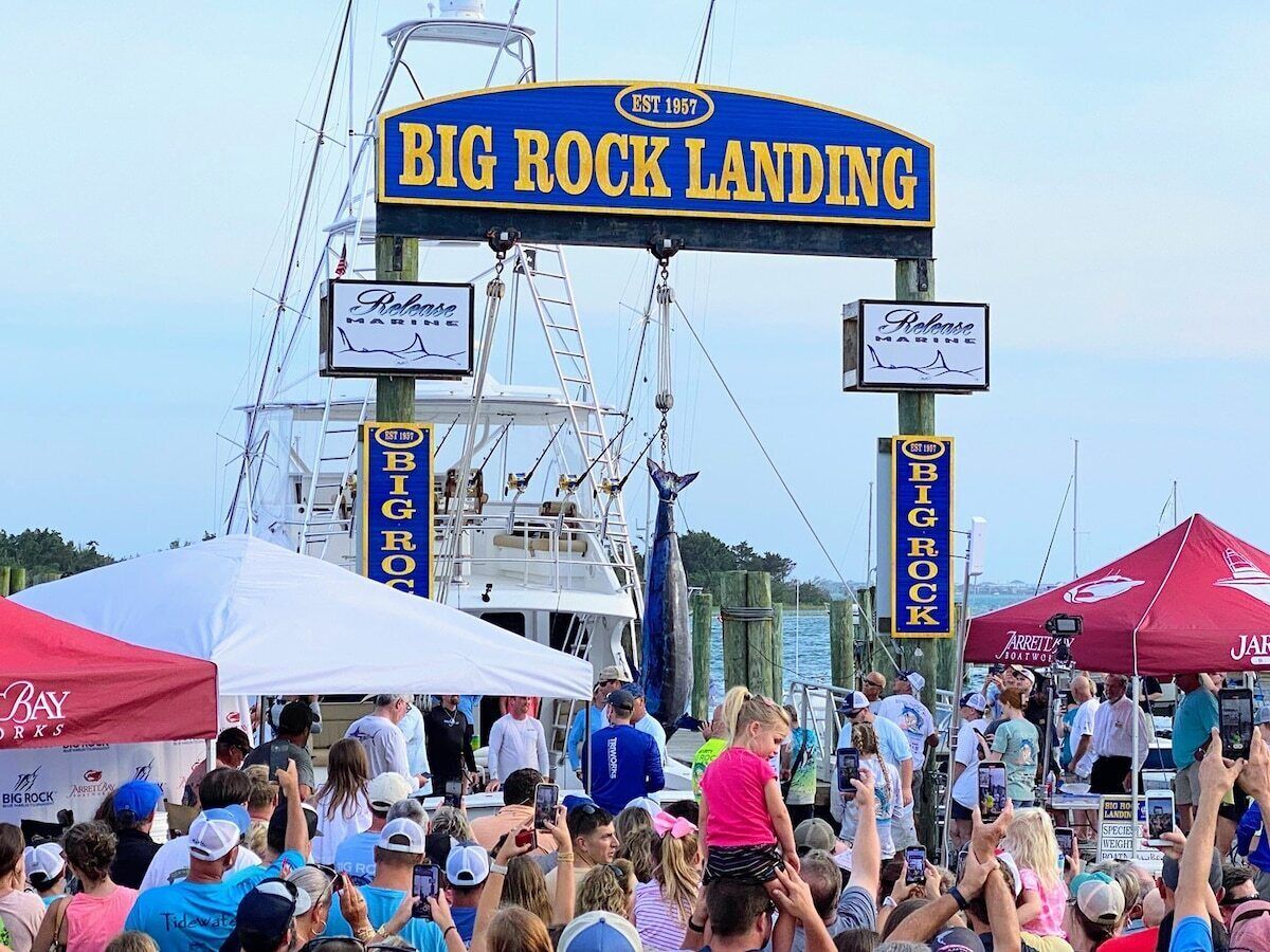 A crowd at a boat launch event with a large sign reading 'BIG ROCK LANDING' and a boat in the background.